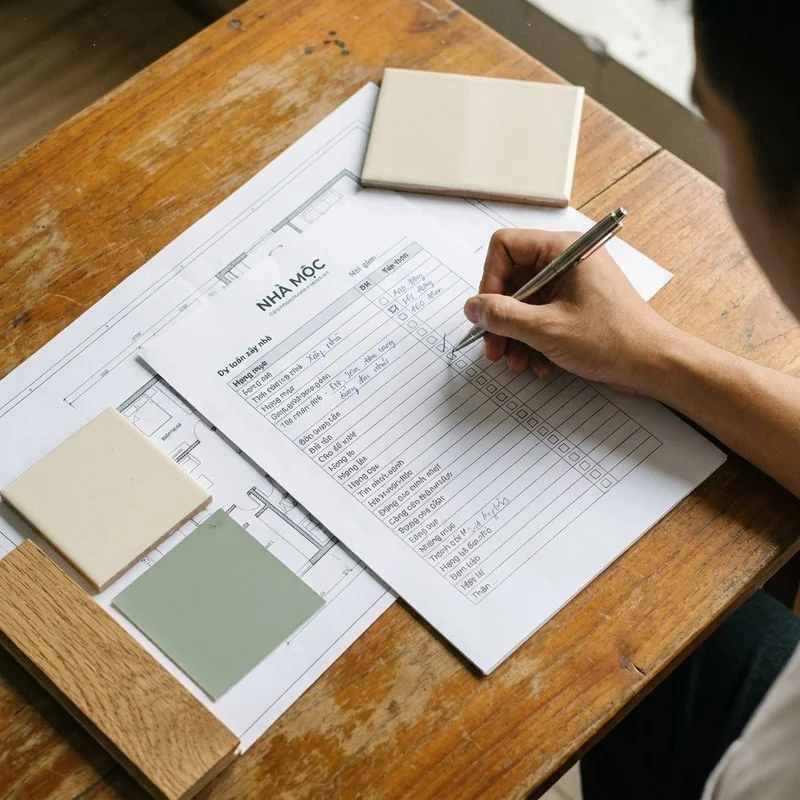 Ảnh minh hoạ: bàn tay đánh dấu checklist hạng mục xây nhà bên bản vẽ mặt bằng nhà phố